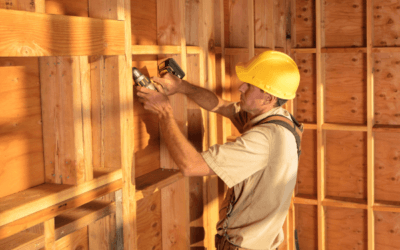 A construction worker wearing a yellow hard hat and tool belt is using a drill to secure wooden framing inside a partially built home. The sunlight casts a warm glow on the exposed wooden structure, highlighting the framework and construction process.