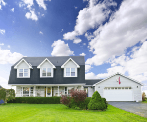 Beautiful two-story home with a well-manicured green lawn, attached garage, and a spacious front porch under a bright blue sky. A residential home for sale in a suburban neighborhood