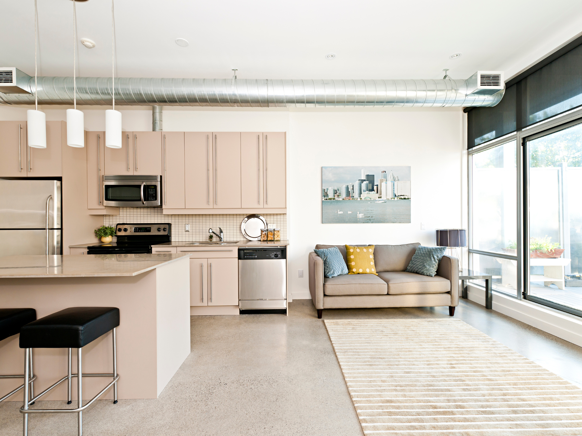 Modern open-concept kitchen and living area inside a West Fargo townhome at The Lights, featuring industrial ductwork, neutral cabinetry, stainless steel appliances, large windows, and a cozy sofa with accent pillows.