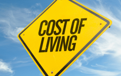 Yellow caution-style road sign with the words "COST OF LIVING" against a bright blue sky, symbolizing awareness of living expenses.